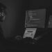 Black and white image of a man looking at a computer set up of three screens with lines of code on one of them. He wears headphones.