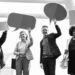 Black and white image of four people each wearing smart clothing and holding up speech bubble signs