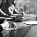 Black and white image of several people sat at a table with notepads and pens and their heads are turned to look at someone speaking