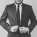 Black and white image of a person in a dark suit and tie, pulling the blazer across themselves